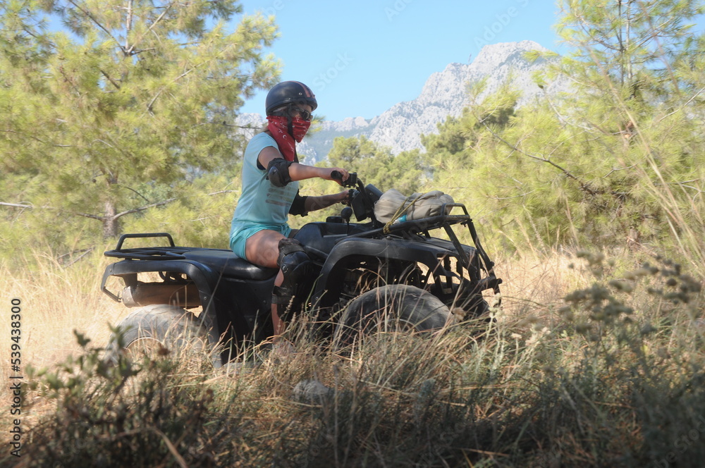 Photo of a girl on a quad bike in a helmet, ammunition. Beautiful landscape, mountain background. FeMale driver riding atv buggy at savannah and rough terrain, safari extreme fun. face not visible. 