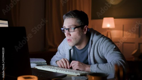 male freelancer in eyeglasses remote works at home on laptop at night darkness. programmer gawp intently on monitor with whole wide eyes. he looks worried and nervous, dissatisfied photo