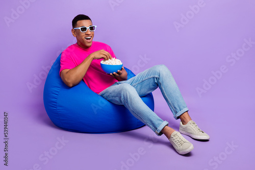 Full size portrait of overjoyed positive man sit comfy bag eat popcorn watch 3d movie isolated on purple color background