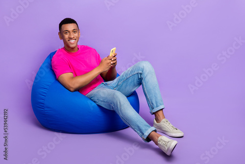 Full length portrait of cheerful person sit comfort bag hold use telephone isolated on purple color background