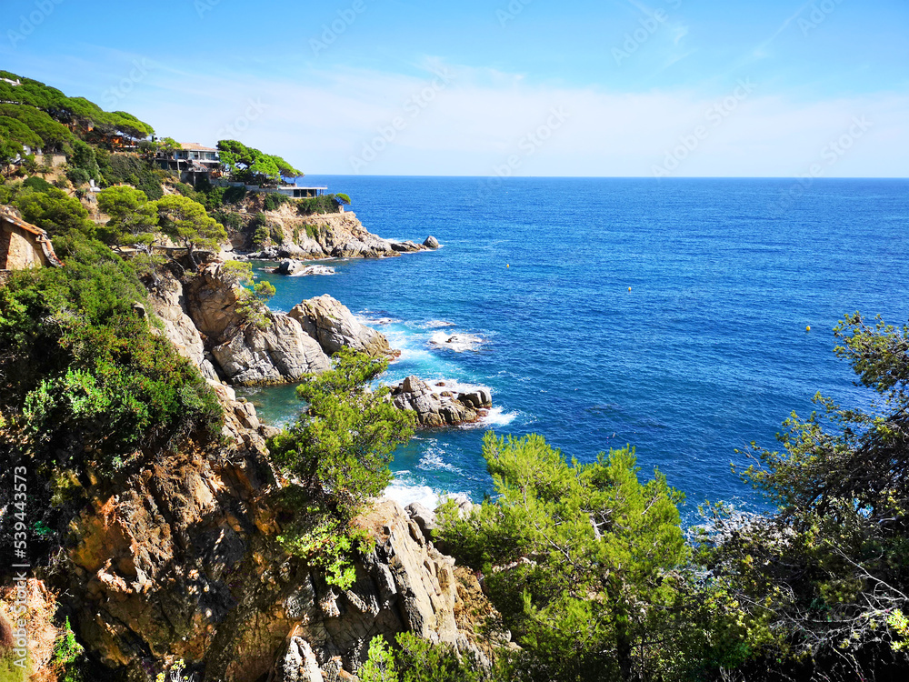 Panoramic photo of the rocky seashore with the sky, with green plants, trees and private houses. The Spanish coast of the Costa Brava, the Mediterranean Sea. Tourism, living in a beautiful place.