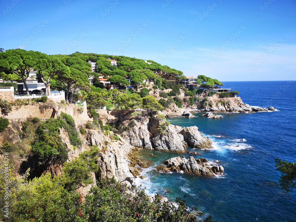 Panoramic photo of the rocky seashore with the sky, with green plants, trees and private houses. The Spanish coast of the Costa Brava, the Mediterranean Sea. Tourism, living in a beautiful place.