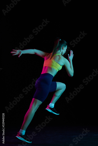 Professional female athlete running isolated on dark background in neon light. Healthy lifestyle, skills, sport, fitness, speed and energy concept.
