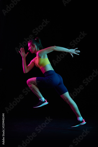 Dynamic portrait of young sportive woman training, running isolated on dark background in neon light. Competition, sport, fitness, speed and energy concept.
