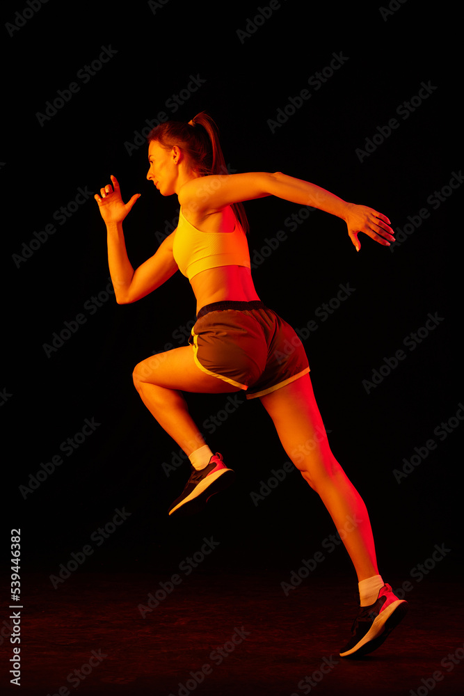 Professional female athlete running isolated on dark background in neon light. Healthy lifestyle, skills, sport, fitness, speed and energy concept.