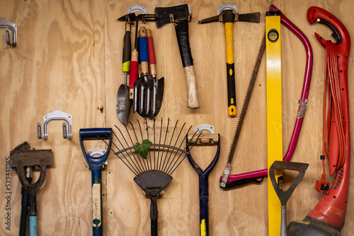 Assortment of DIY gardening tools and equipment hanging organised on wooden wall inside garden shed. 