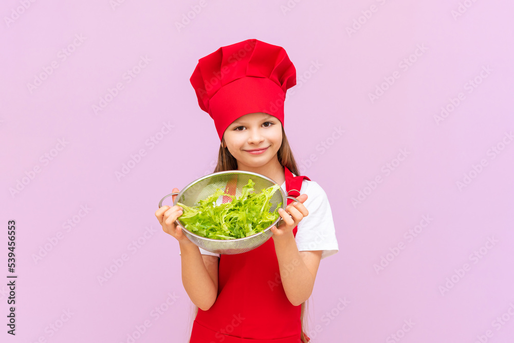 a smiling girl in a red chef's suit holds a sieve with a fresh green salad in her hands. a smiling girl in a red chef's suit holds a sieve with a fresh green salad in her hands.