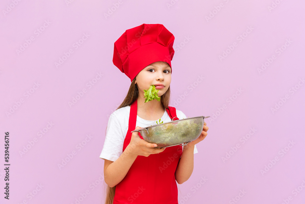 A little beautiful girl in a red chef's costume is eating a fresh salad. The child loves to cook at home.