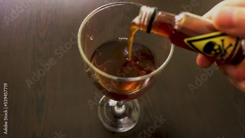 male hand pouring a poison or  a drug from a small bottle into a glass with an alcoholic liquid for drinking - toxic symbol of a skull, mixed with red wine - dangerous concoction for a poisoning