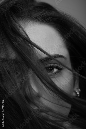 Close-up beautiful woman with seductive look studio portrait. Model covering part of her face with hairs by blowing wind. Mysterious and sensual look. Eye is in camera focus. Black and white image