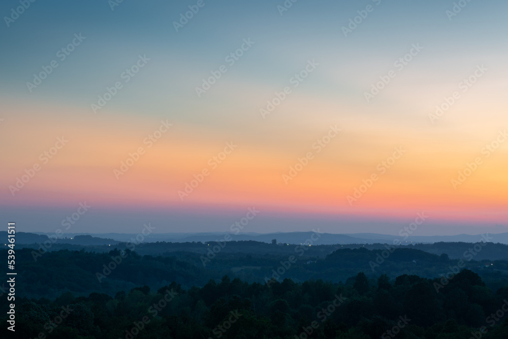 Hill layers separated with haze and vibrant glow in sky above horizon at twilight, hilly rural landscape with last light on clear sky
