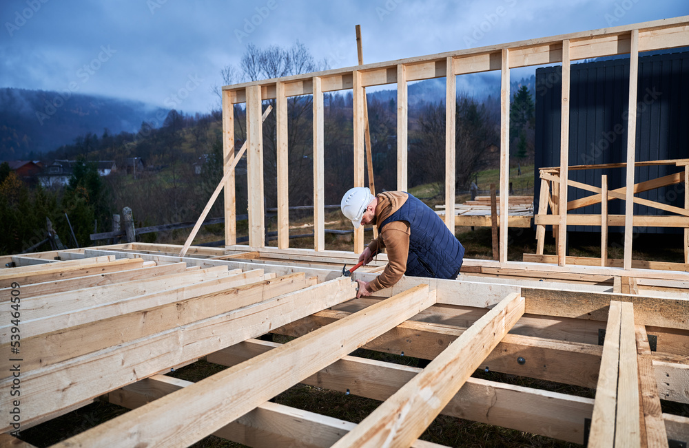 Man worker building wooden frame house on pile foundation. Carpenter hammering nail into wooden plank, using hammer. Carpentry concept. Man worker building wooden frame house on pile foundation. Carpenter hammering nail into wooden plank, using hammer. Carpentry concept.