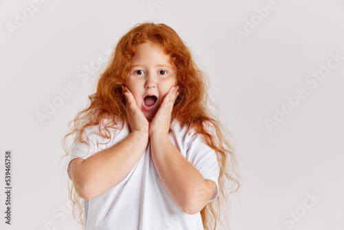 Shock, surprise. Cute little girl, kid with long curly red hair looking at camera isolated over white background. Concept of children positive emotions, beauty
