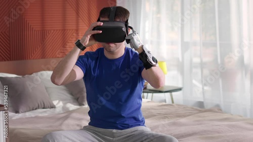 Young man with a bionic prosthetic arm wearing virtual reality glasses sitting on bed  photo
