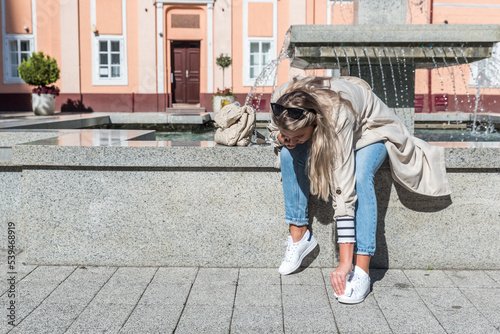 Fotografie Young woman sitting at the town square on the water fountain cleaning and rubbing dog or pigeon poop from her new white shoe