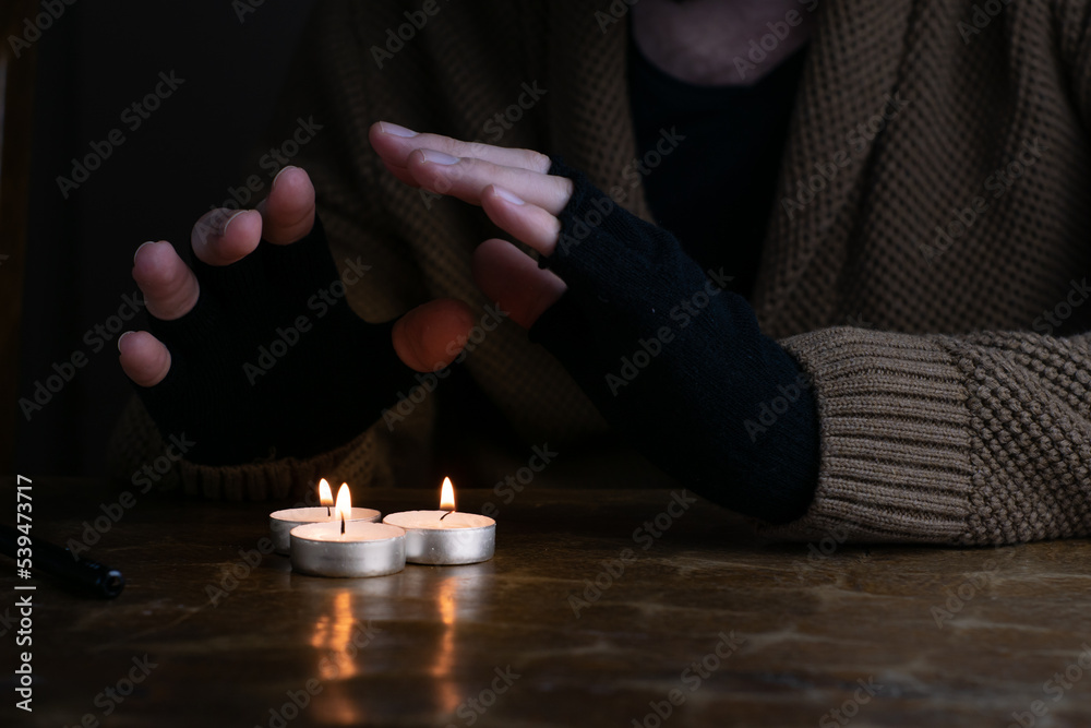 Man in sweater and fingerless gloves warming up his hands with tealight candles during energetic crisis in Europe.