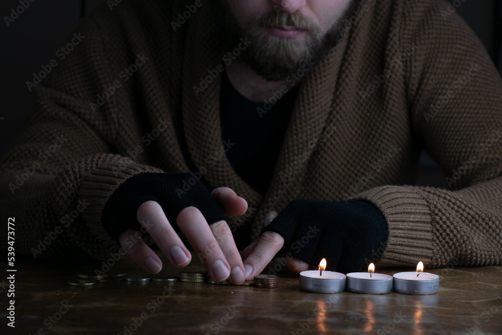 European poor bearded man counting euro coins during energy crisis in Europe. A worried man in warm sweater and gloves counts money in candlelight.