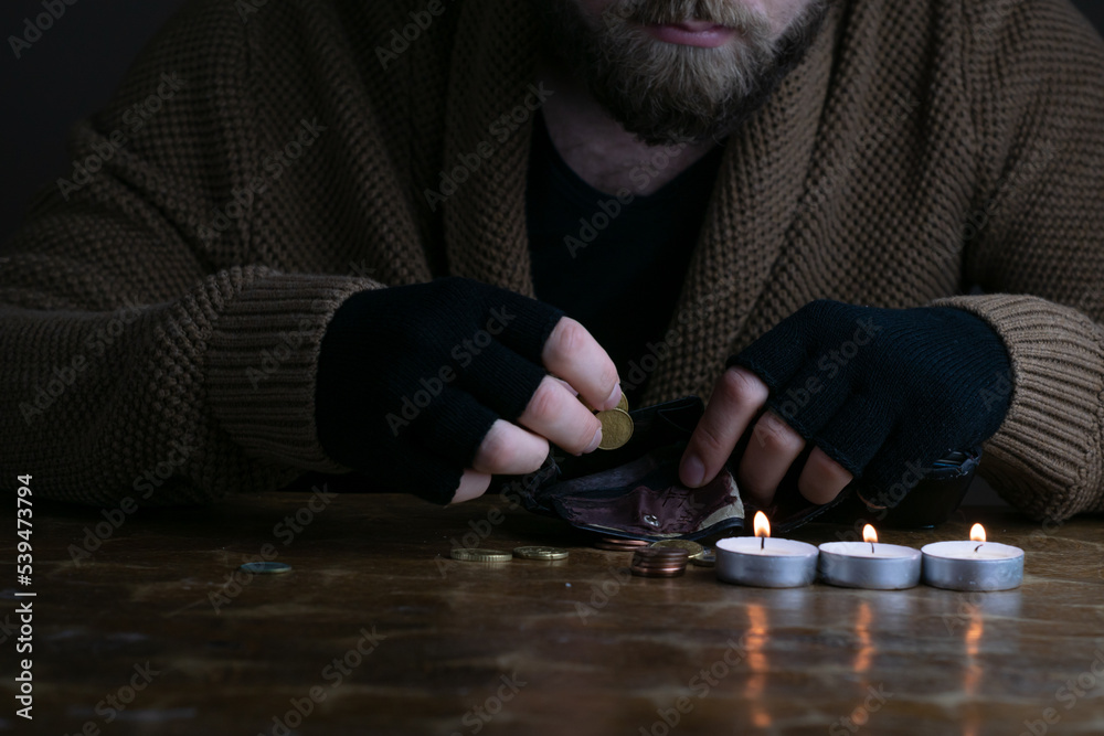 European poor bearded man counting euro coins during energy crisis in Europe. A worried man in warm sweater and gloves counts money in candlelight.