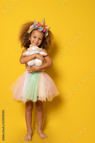 Cute child girl in elegant tulle dress and unicorn headband celebrates birthday party holding teddy bear over yellow background.