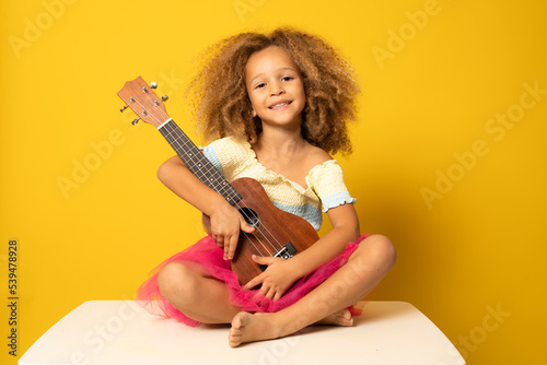 Cute little girl in summer clothing playing ukulele over yellow background. Happy vacation concept.