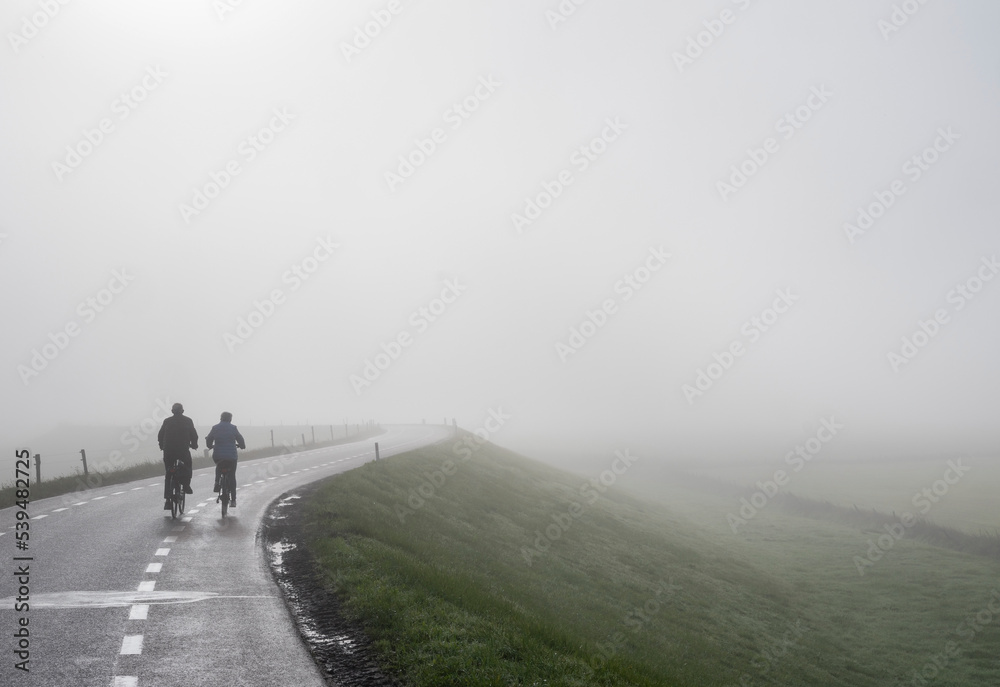 Obraz premium couple on bicycle rides in mist on dike near river rhine in holland