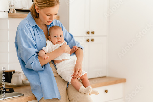 Young white mother wearing shirt holding her baby in arms at home