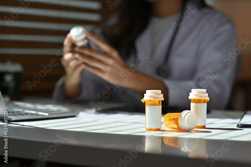 Selective focus on pill bottles with empty label and female pharmacist checking pill bottles and reading drug labels as the background.