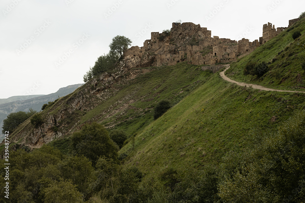 Ruin of old stone castle on peak of in mountain in Dagestan with road on green slope, majestic mountains landscape, canyon in highlands in overcast. Historic adventure, tourist trip.