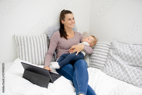 pretty young mother sitting, lying in bed, working on notebook and holding her sleeping baby in her arms