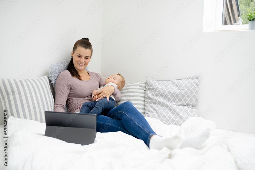 pretty young mother sitting, lying in bed, working on notebook and holding her sleeping baby in her arms pretty young mother sitting, lying in bed, working on notebook and holding her sleeping baby in her arms