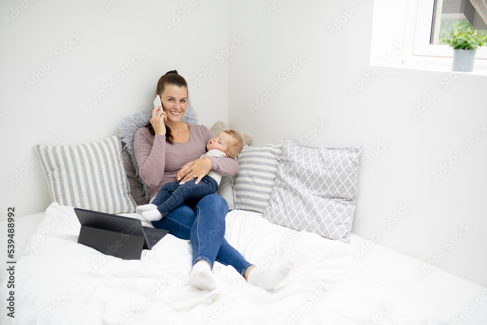 pretty young mother sitting, lying in bed, working on notebook and holding her sleeping baby in her arms pretty young mother sitting, lying in bed, working on notebook and holding her sleeping baby in her arms