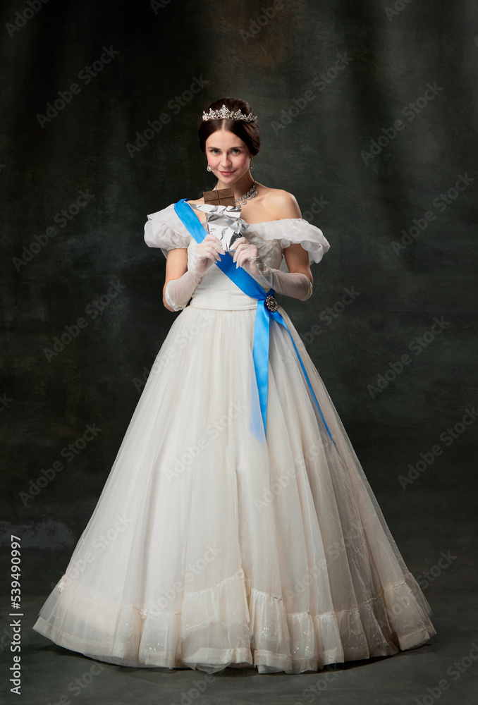 Young queen. Portrait of adorable girl in image of medieval royal person in renaissance style dress isolated on dark background. Comparison of eras, beauty, history, art Young queen. Portrait of adorable girl in image of medieval royal person in renaissance style dress isolated on dark background. Comparison of eras, beauty, history, art