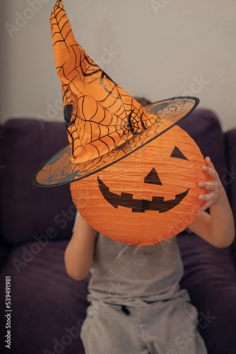 Boy holding artificial pumpkin in orange hat at head level, halloween concept. High quality photo