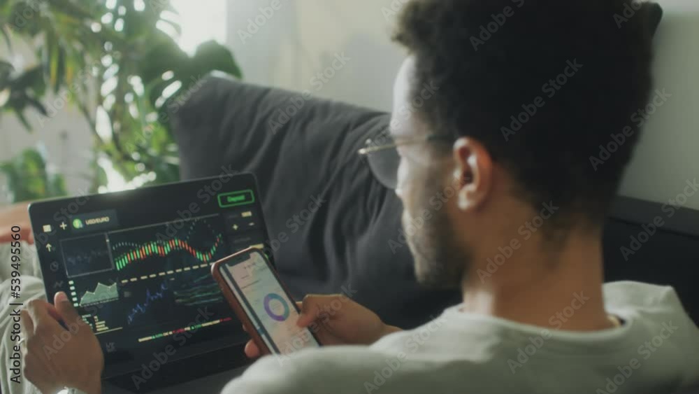 Over the shoulder shot of young man sitting on sofa at home, analyzing stock market graph on laptop and using finance app on phone while trading online