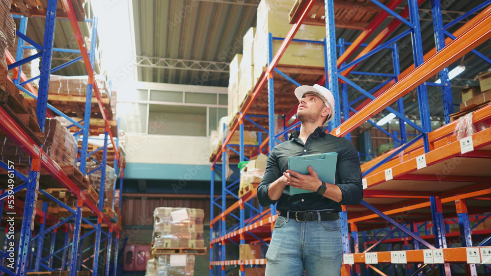 Caucasian logistics employee is examining the stock of cargo cardboard boxes on shelves and recording on paperwork. Young adult logistics manager concentrate on working to manage the stock of goods. Caucasian logistics employee is examining the stock of cargo cardboard boxes on shelves and recording on paperwork. Young adult logistics manager concentrate on working to manage the stock of goods.