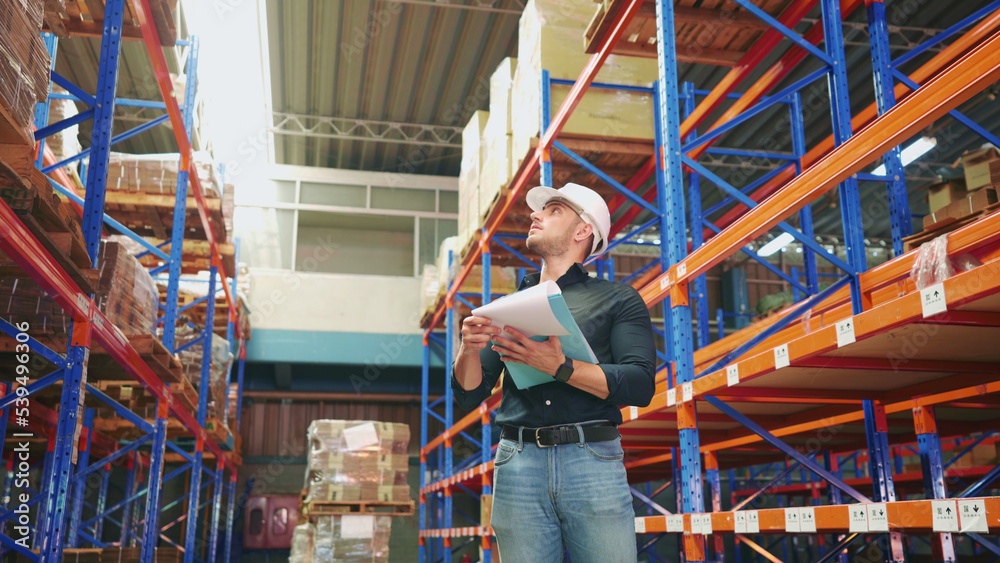 Caucasian logistics employee is looking at shelves and counting number of cargo cardboard boxes to record on paperwork. Logistics manager is examining the stock of goods being prepared for shipping. Caucasian logistics employee is looking at shelves and counting number of cargo cardboard boxes to record on paperwork. Logistics manager is examining the stock of goods being prepared for shipping.