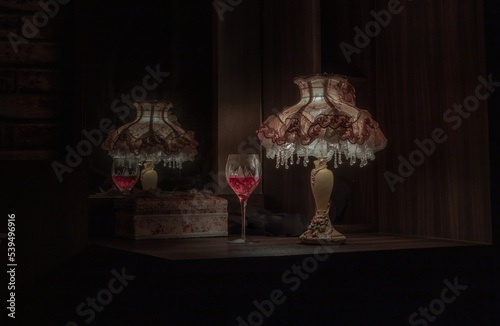 A glass of red wine puts on dressing table in the dim light of Vintage night lamp with reflection from mirror in the bedroom at night. Light and Shadow, Dark tone, Selective focus.