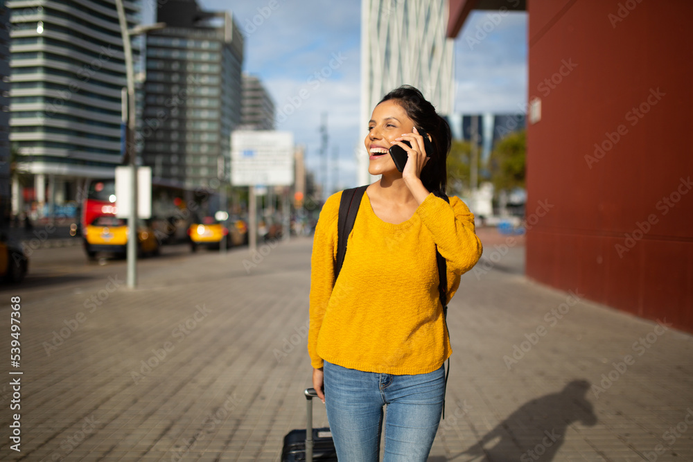 smiling young woman walking with suitcase and talking with phone