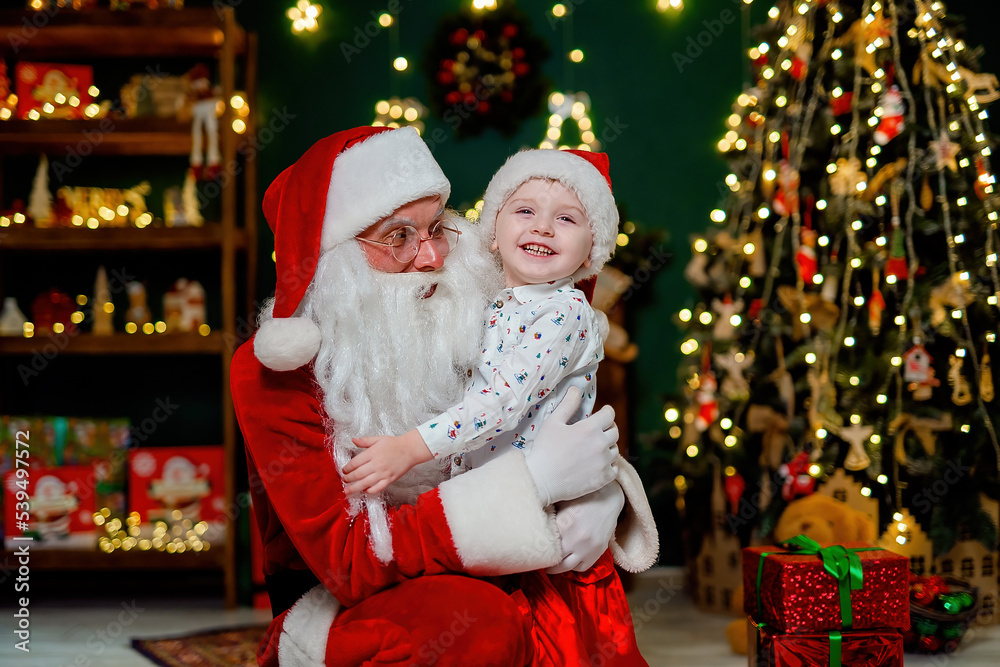 Laughing little boy in Santa's hat hugging with Santa Claus near Christmas tree. New Year concept Laughing little boy in Santa's hat hugging with Santa Claus near Christmas tree. New Year concept