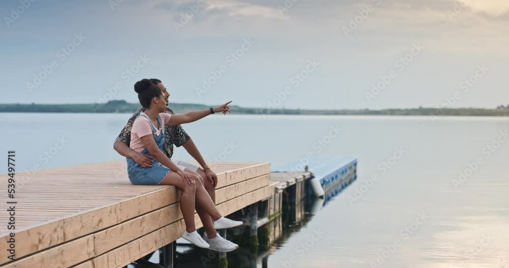 Side view of happy African American family couple sitting on dock looking at lake with beautiful sunset spending time together. Husband and wife resting on jetty on summer. Weekend rest