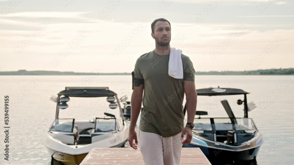 Portrait of African American man in shirt and shorts with towel on shoulder walking on dock between modern boats on lake. Handsome young male on pier outdoor near yachts on water. Sport and recreation
