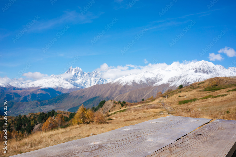 Close-up of an empty wooden bench against a blurred background. Edge of table on a sunny day in the mountains. Snow covered mountains and a blue sky. hike, break, view