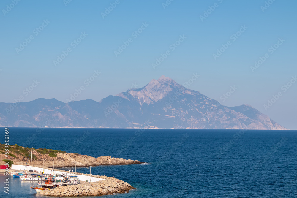 Scenic morning view of holy Mount Athos seen from small harbour port Sarti on peninsula Sithonia, Chalkidiki (Halkidiki), Greece, Europe. Summer vacation at Aegean Mediterranean Sea. Fishermen boats