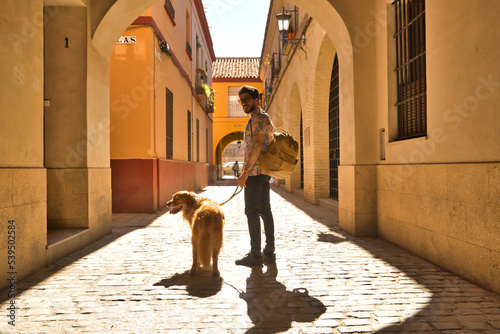 Young Hispanic man with beard and sunglasses with his dog holding on to the leash walking along a sunny street at sunset. Concept animals, dogs, love, pets, golden.