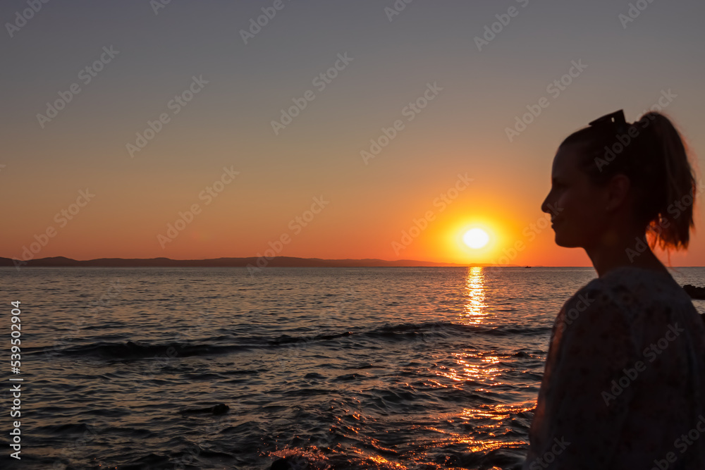 Fototapeta premium Silhouette of tourist woman holding the sun with panoramic view of sunset over Aegean Mediterranean Sea on Karydi beach, peninsula Sithonia, Chalkidiki (Halkidiki), Greece, Europe. Romantic atmosphere