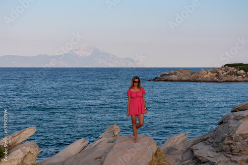 Beautiful woman in red dress standing on rock with panoramic view of Mount Athos seen from Karydi beach, Sithonia, Chalkidiki (Halkidiki), Greece, Europe. Summer vacation at Aegean Mediterranean Sea