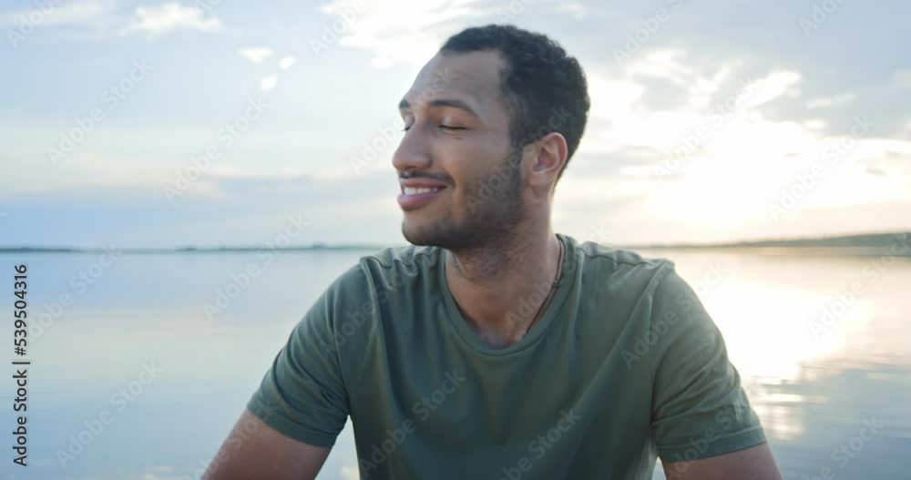 Close up shot of joyful young man in good mood resting on lake looking at camera and smiling on sunset recording. Cute African American happy male on nature outdoors alone with water on background