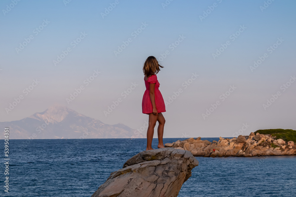Beautiful woman in red dress standing on rock with panoramic view of Mount Athos seen from Karydi beach, Sithonia, Chalkidiki (Halkidiki), Greece, Europe. Summer vacation at Aegean Mediterranean Sea