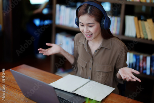 Portrait of a smiling Asian teenage girl wearing headphones and using a computer for online video conferencing in a library