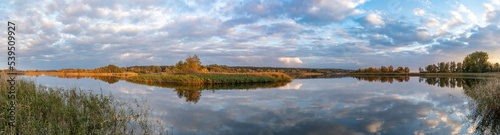 Landschaft mit Bäumen, Schilf, wolkigem Himmel und der Spiegelung der Wolken im See